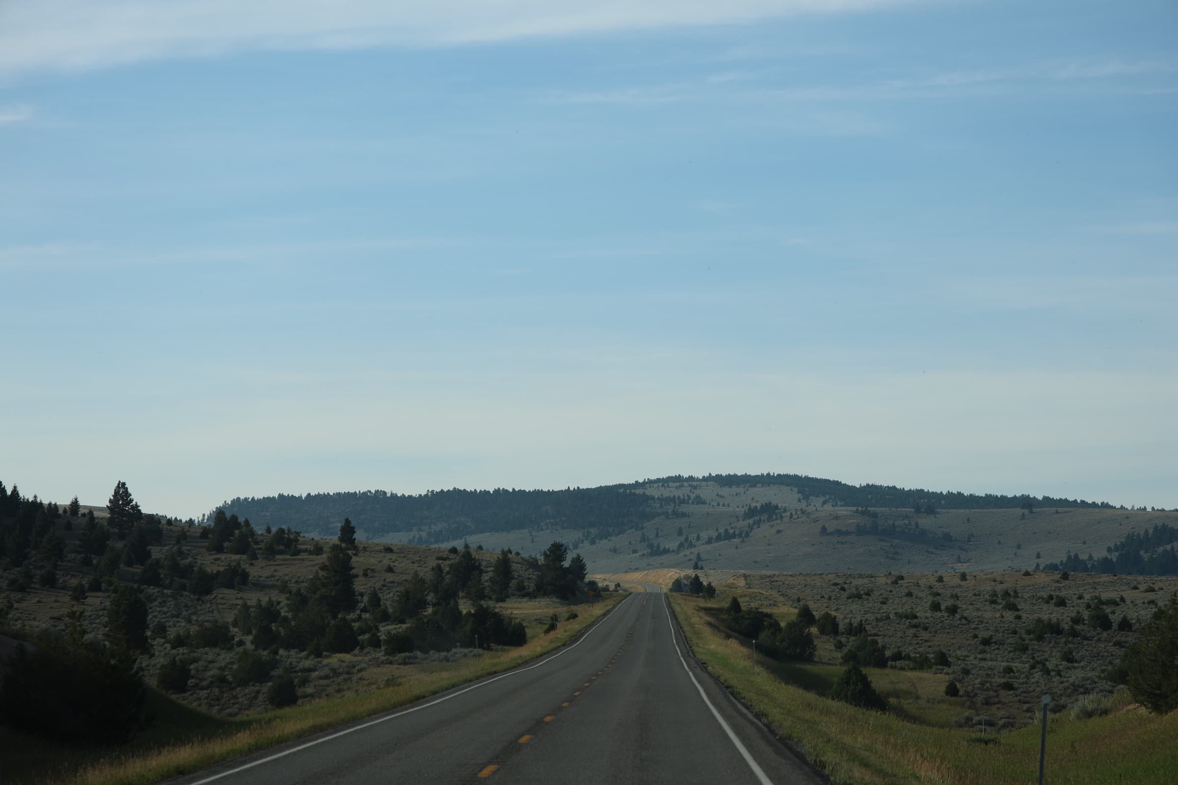 An empty two-lane highway running out across Montana rangeland under a wide, pale sky.